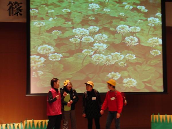 スクリーンには花の絵が写り、舞台では女性達ががランドセルに黄色い帽子を被り劇をしている写真